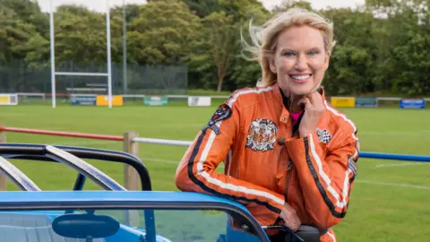 Channel 5 A blonde Anneka Rice in a bright orange leather jacket. She is resting her elbow on the edge of her blue and yellow car. A rugby pitch is in the background.