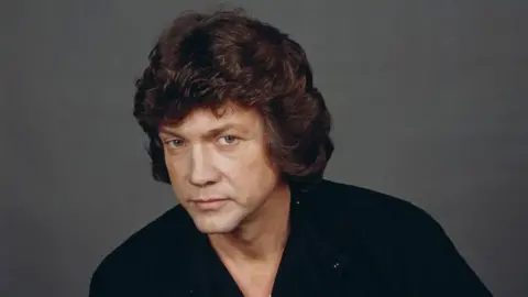 Michael Putland/Getty Images A man with curly brown hair is looking at the camera. He is wearing a dark top.