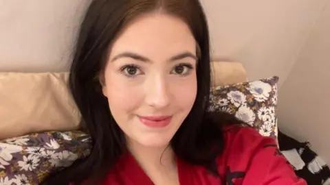 Holly Harris Holly has shoulder length dark hair and brown eyes and is sitting down. She is wearing a red top.