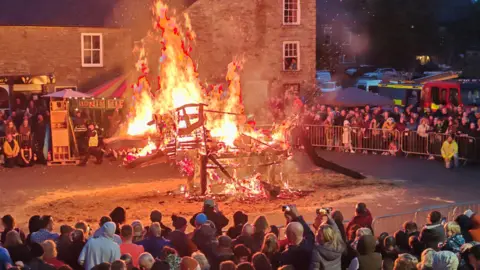 Adrian Ashurst A wooden structure in the shape of a wolf which is covered in fire. It is standing in a village square on top of a pile of sand. Barricades form a circle around the fire, with crowds of people watching on. A fire engine can be seen parked in the background.