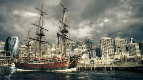 Getty Images A three masted wooden sailing ship, about 100ft long, is docked in the harbour of a modern city, with tall office buildings in the background under a grey sky.