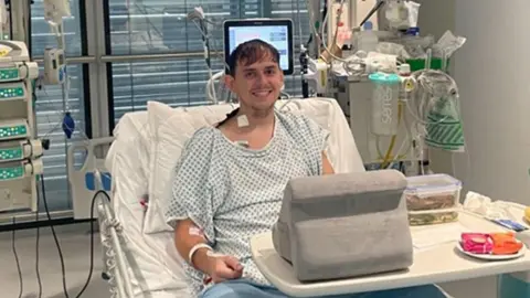 The PHA Group/PA Wire A man sits smiling in a hospital bed surrounded by tubes, wires and medical machines.