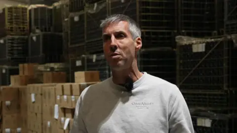 Sam Lindo from Camel Valley Vineyard stands inside a warehouse where boxes of wine made at the vineyard are stored. He is stood next to two metal poles. His is gesturing with his hands out stretched. He is wearing a grey jumper with the company's logo on it.