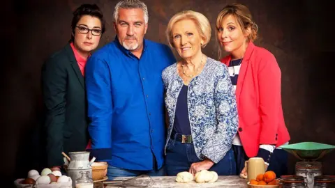 Love Productions/BBC Sue Perkins, Paul Hollywood, Mary Berry and Mel Giedroyc, standing around a table, with baking ingredients on it, they are all looking at the camera. 