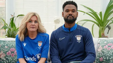 BBC A woman with long blonde hair, wearing a blue top sits next to a man with dark hair and a dark beard, wearing a darker blue top, on a sofa in front of windows with closed Venetian blinds.