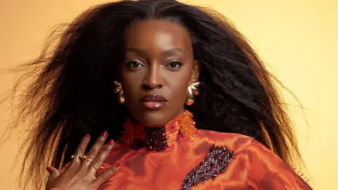 Coco Sarel Woman wearing high neck orange top and pouting at the camera 