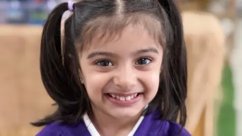 BBC A young child smiles at the camera  with long black hair, pulled up in ponytails either side of her head. She wears a purple top.