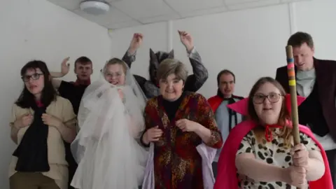 BBC A group of people dressed up in spooky costumes, including a ghost and a werewolf. They are all smiling, some with their hands in the air.
