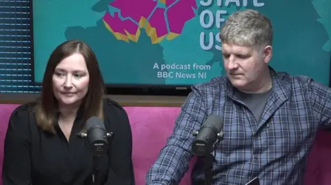 BBC A screen shot of the Brysons filming a podcast. They are sitting in front of black microphones and in front of podcast promo posters. Ms Bryson has long brown hair and is wearing a black blouse. Mr Bryson has grey hair and is wearing a blue checked shirt.
