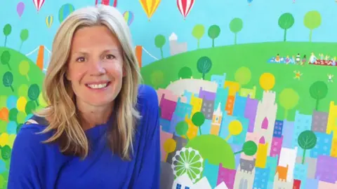 Jenny Urquhart Wearing a blue top, Jenny smiles at the camera whilst posing in front of one of her artworks. She has blonde hair hanging loose below her shoulders. The art depicts hot air balloons flying over Bristol. The Clifton Suspension Bridge can be seen in the work behind her, as can children playing in a park.