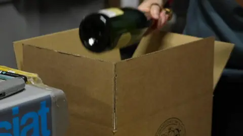 BBC A person places a bottle of win in a cardboard box in a warehouse. A grey machine with "siat" written on it in blue letters is next to the box. A grey Stanley knife is on top of the machine.