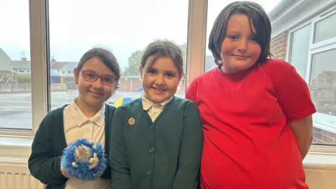 BBC Three children stand in front of a window with their school playground in the background outside. Scarlett stands to the left wearing a white polo top and green cardigan. She's smiling at the camera holding her blue hedgehog teddy bear. Luna is in the middle with the same uniform and Bailey is to the right wearing a red sports t-shirt.