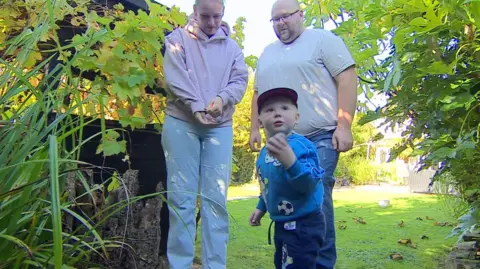 BBC / Peter Whittlesea A boy wearing a blue jumper with football print patterns. Behind him is a woman wearing a light link hoodie and jeans and a man in a light grey shirt and black trousers. They are standing in a garden.