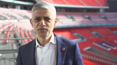 Harry Low / BBC Sadiq Khan stood in the stands at Wembley Stadium. He is wearing a white shirt with an unbuttoned collar and a blue jacket, with a straight face looking at the camera.