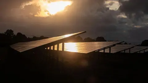 Getty Images Rows of solar panels shrouded in darkness. An orange glow shines through a crack in the clouds and reflects off the panels.