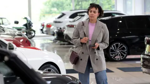 Getty Images Woman wearing long grey blazer and jeans standing in a car showroom with various cars lined up