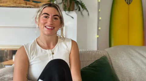 BBC Laura has blonde hair tied back sits on a sofa, smiling. Behind her are two yellow surfboards and a small flag on the wall. The setting appears relaxed and homey.