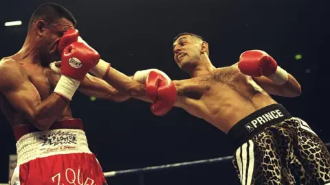 Getty Images A boxer in red trunks labelled "Vazquez" taking a punch to the face from an opponent in leopard-print trunks labelled "Prince" during a match in a boxing ring.