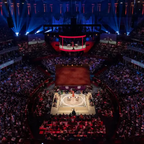 Getty Images A view of the Royal Albert Hall, showing the crowds surrounding the ring, which has two sumos fighting in it, with the judge looking over them. Above that is the temple roof, which has tassles hanging down, and above that is the circular LED screen which has the match playing on it