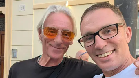 Phil James Singer Paul Weller is dressed in a black t-shirt and orange sunglasses. He has long white hair and has his arm around a man in a white-shirt and black glasses. They are stood outside a cream building with a wooden door.