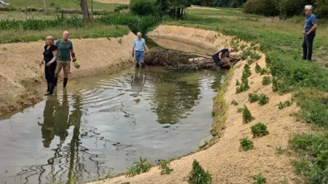 HMWT Four people, standing by a river, with a bank and foliage on it, with a tree in the middle of the river. There is grass around the water and trees. 
