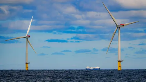 PA Media A ship passing between two offshore wind turbines on a dark blue sea under a light blue sky with grey and white clouds.