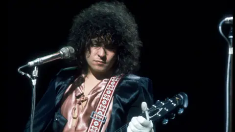 BBC A man with long, dark, curly hair stands in front of a microphone with a guitar 