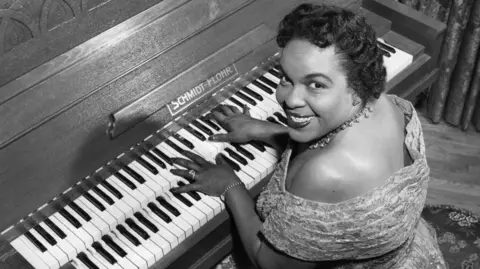 BBC Winifred Atwell smiles at camera whilst her hands are on the piano. 