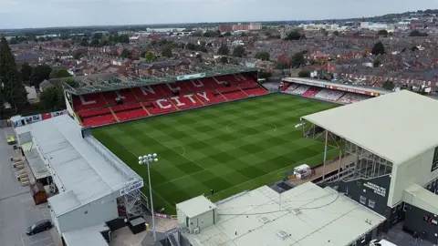 BBC Aerial view of Sincil bank the home ground of Lincoln City. It has four stands and floodlights with the city of Lincoln in the background. The main stand has white seats spelling out Lincoln City on the red seat around it.
