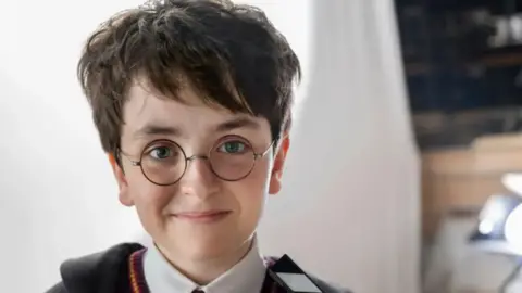 Aidan Monaghan/HBO The image shows a young boy dressed in a costume from the Harry Potter series. He is wearing a white shirt, and a black robe with red and gold edged jumper. He is wearing round glasses and has short, dark brown hair.