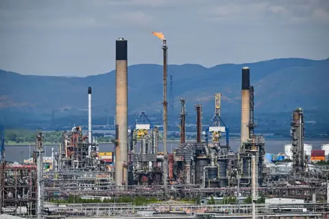 Getty Images Grangemouth oil refinery stands in front of the Clyde river with mountains in the background. The refinery has several tall towers, with one emitting flames