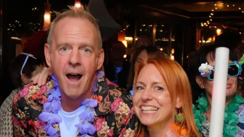 Sussex Partnership NHS Trust The DJ Fatboy Slim, wearing a floral short-sleeve shirt over a white t-shirt with a garland of flowers around his neck, stands beside a woman with red hair and in front of two people also dressed in bright clothing and wearing sunglasses.