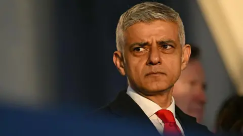 WPA Pool/Getty Images Sadiq Khan facing the camera head-on, looking into the distance away from the camera. He is wearing a black suit with a red tie.