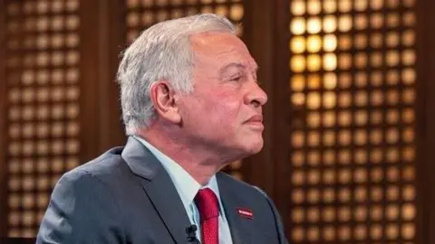 BBC Jordan's King Abdullah, wearing a black suit jacket, pale blue shirt and red tie, pictured in profile