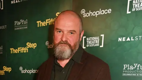 Getty Images A man with a grey beard and shaved head wearing a brown jacket and green shirt. He standing in front of a poster that reads "The Fifth Step, @sohoplace, National Theatre of Scotland." He has a slight smile.