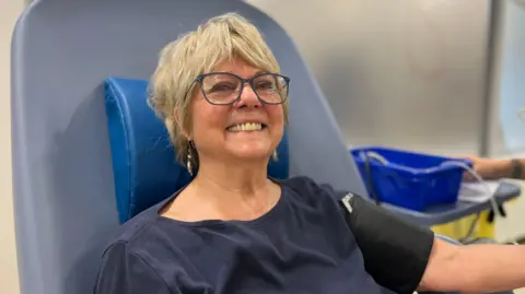 George Carden/BBC Mo Parker sat in a chair about get her blood taken. She has a blood pressure monitor on her arm and has blond hair and glasses.