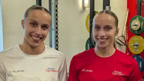 University of Essex Abigail and Emily Roper at the gym with large weights behind them. They have their blonde hair in ponytails and are smiling. Abigail is wearing a white T-shirt and Emily a red one.