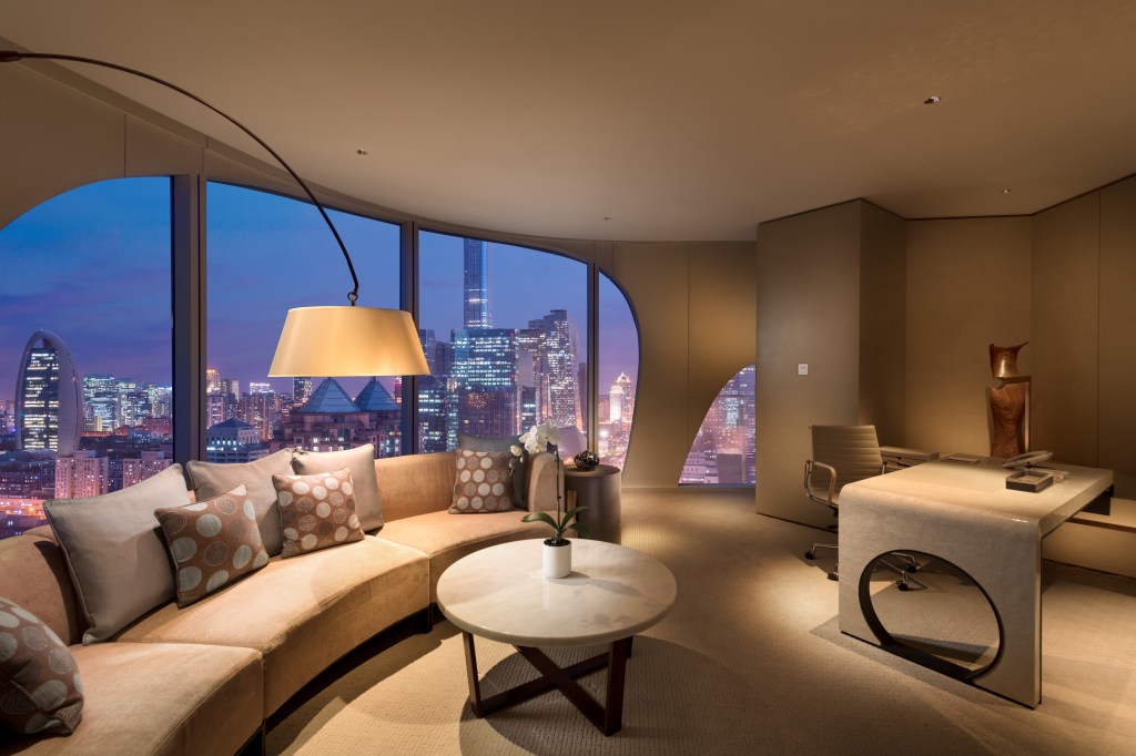 A living space with curved couch with window behind looking out over Beijing, and desk
