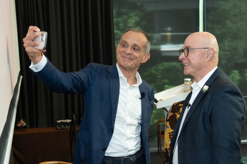 Two men stand side by side on stage at an event. One is taking a selfie while they both smile for the camera. 