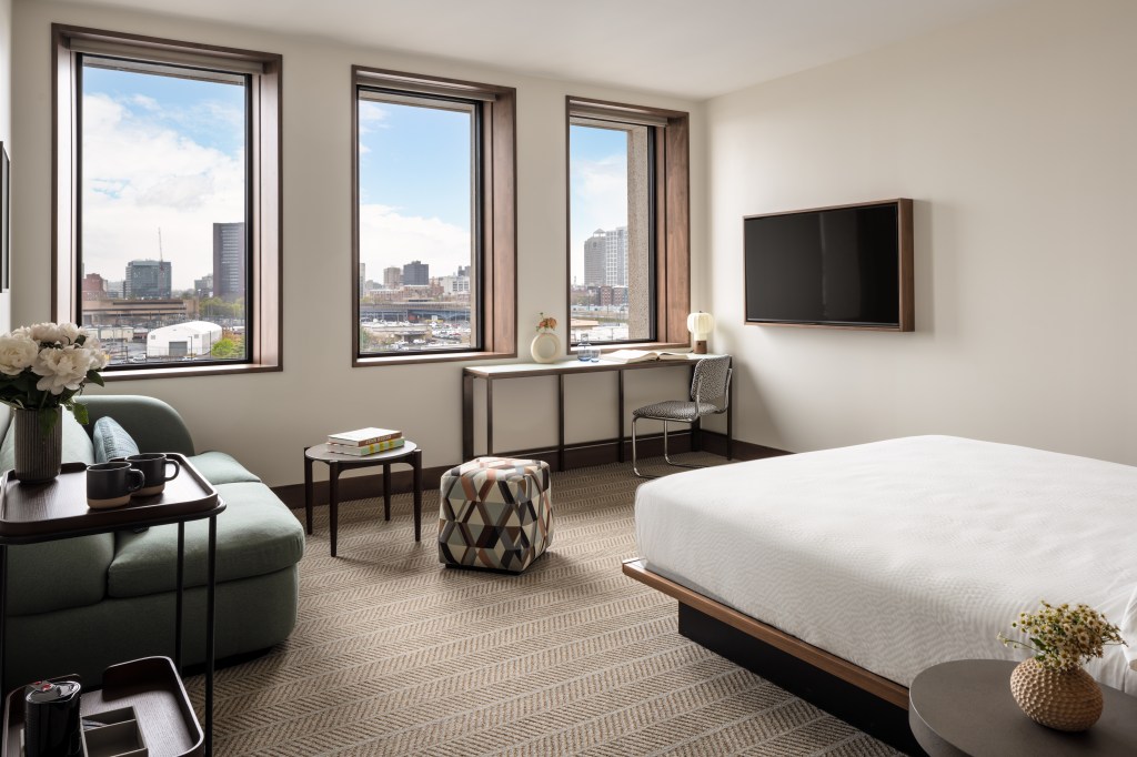 A bright bedroom with bed, couch and desk, and windows looking out at the city.