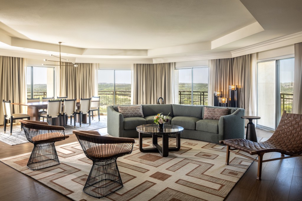 A living space with couch, chairs, and dining table, with many windows overlooking the balcony and the view behind.