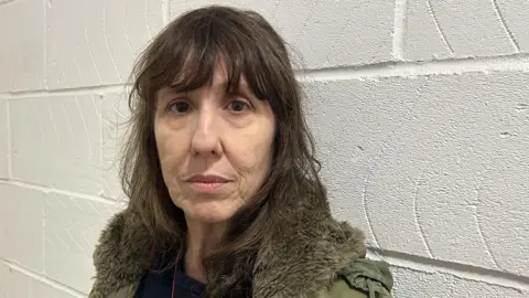 BBC A woman with long dark brown hair. She is wearing a green coat and looking at the camera