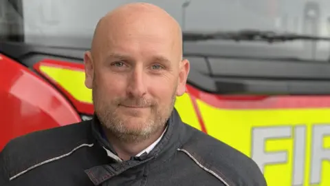 Cambridgeshire Fire and Rescue Service Paul Whitaker is in his uniform, standing in front of a red and yellow fire engine, facing the camera with a composed expression. The word 'FIRE' is partially visible on the vehicle behind, emphasizing the emergency services setting. The firefighter’s dark uniform, detailed with light stitching, reflects the seriousness and dedication of the profession."