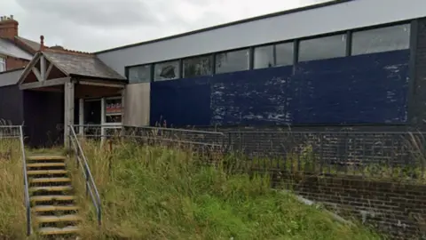 Google The former Stanley library is an empty one-storey building made of brown bricks and surrounded by overgrown grass. 