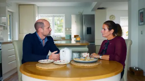 Kensington Palace Prince Williams and Rhian Mannings sat around her kitchen table talking with a cup of tea. On the table there is a tea pot, two heart patterned plates and a plate of Welsh cakes. Williams, on the left, wears a navy quarter zip and a light blue shirt. On the right, Rhian wears a long sleeved burgundy blouse with black bow details. She has dark brown hair in a ponytail.