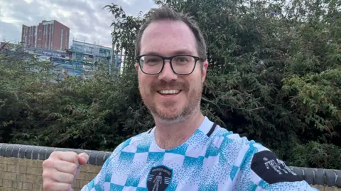BBC/Adam Oxley A young man with brown hair and glasses wears a blue and white football shirt. He is standing in front of some trees with a building in the background and is punching the air with his fist. 