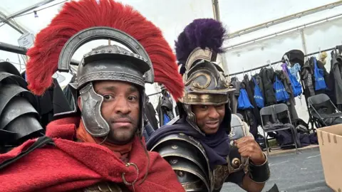 Supplied Two men take a selfie as they sit side by side in a tent which has hanging rails along the perimeters full of coats and bags. They are both wearing Roman Empire-style military costumes.
