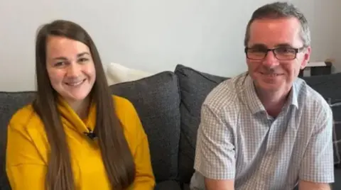 West Midlands Ambulance Service A woman with long brown hair and a yellow long-sleeved top sitting next to a man with short brown hair and chequed short-sleeve shirt and glasses. They are sat on a dark grey sofa, which is against a light grey wall.