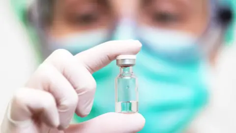 Getty Images A doctor holds up a clear-glass vial his white gloved hand. He wears a green face mask and clear goggles.