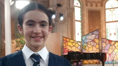 Marcela Arancibia Zoe. She has dark hair tied back, wearing a navy school uniform, white shirt and navy striped tie. She is standing in a church, in the background there are stained glass windows, a black grand piano and studio lights.